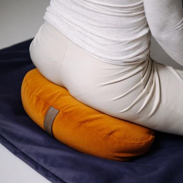 Zbliżenie na siedzącą osobę medytującą na musztardowej poduszce do jogi, relaksacji i medytacji WellB w kształcie fasolki, na granatowej macie.